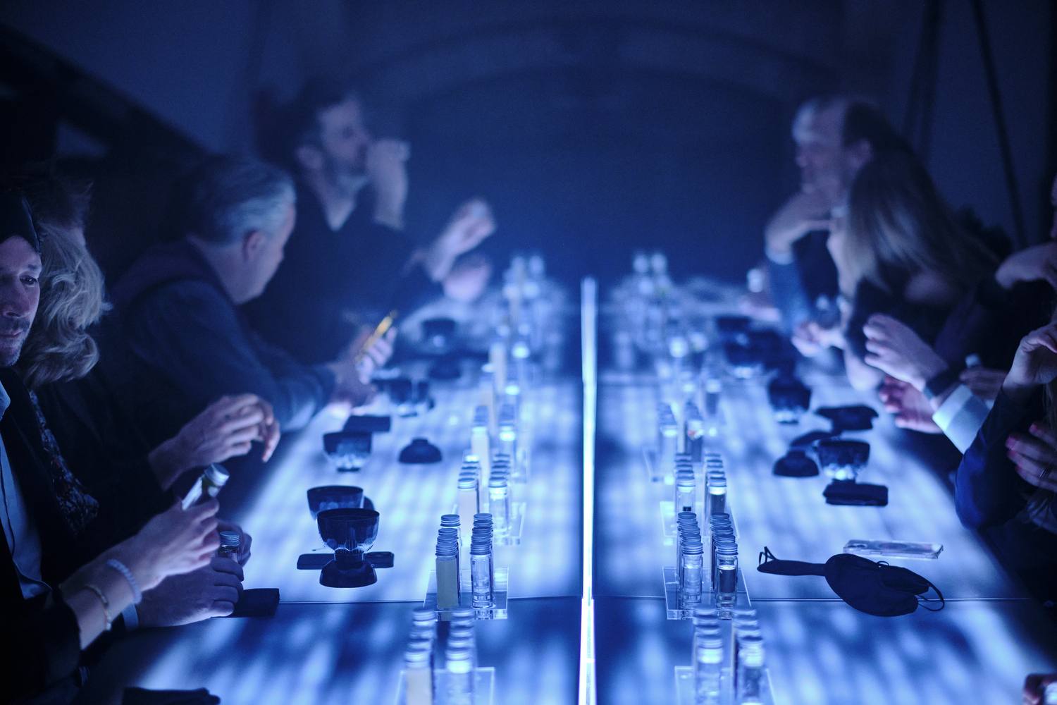 Guests leaning over the illuminated table, tasting from glass vials in the dark